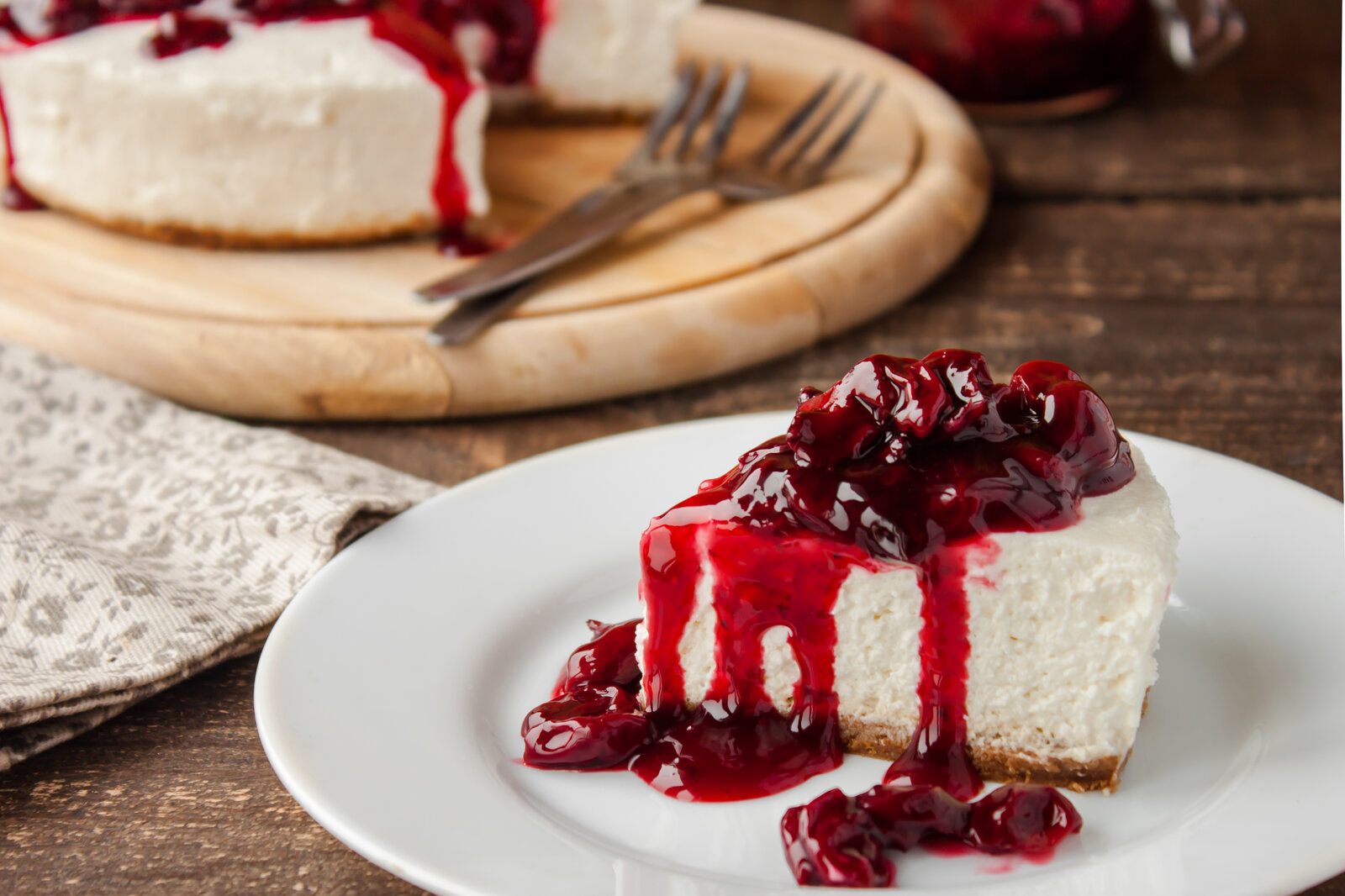 Stück Käsekuchen mit Kirschkompott auf einem Teller, dahinter der gesamte Kuchen. | © Adobe Stock / yuliiaholovchenko