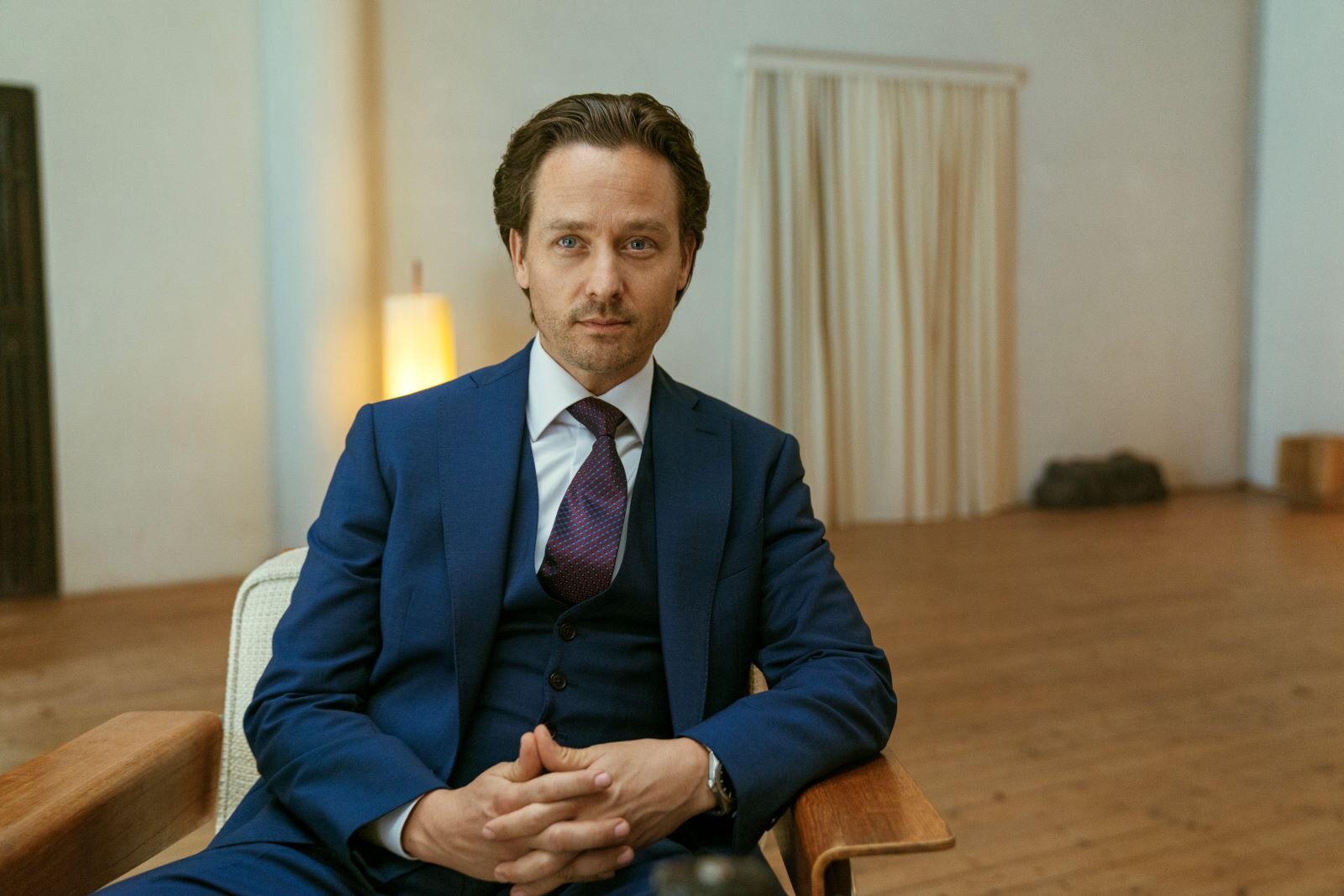 Tom Schilling als Björn Diemel sitzt ernst blickend auf einem Holzstuhl in einem minimalistischen Raum. | © Netflix / Achtsam Morden