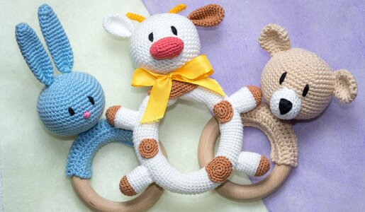 Drei gehäkelte Baby-Greiflinge in Form eines blauen Hasen, einer weißen Kuh mit gelber Schleife und eines beigen Bären auf pastellfarbenem Hintergrund. | © elen31/stock.adobe.com