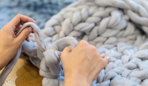 Hände einer Frau flechten hellgraue, dicke Wollstränge auf einem Holztisch, im Hintergrund blaue und rosafarbene Strickstücke. | © Photopixel /stock.adobe.com