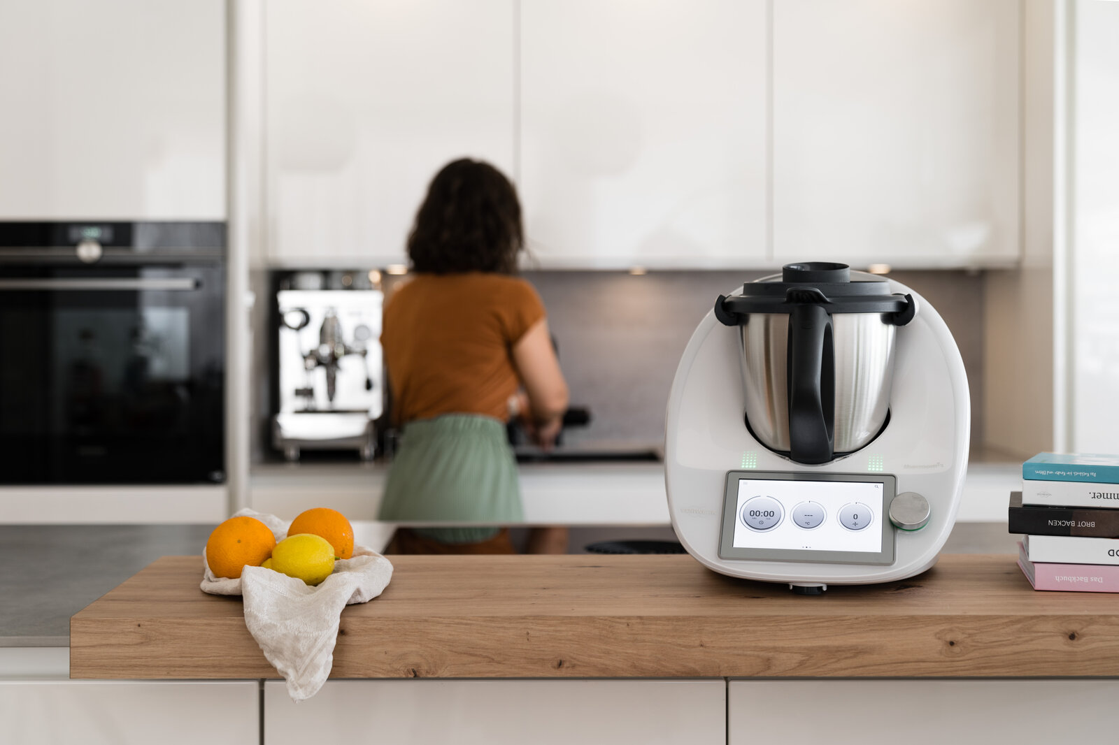 Thermomix auf Holzarbeitsfläche mit Zitrusfrüchten, im Hintergrund kocht eine Frau mit braunen Locken in einer hellen Küche. | © Vorwerk