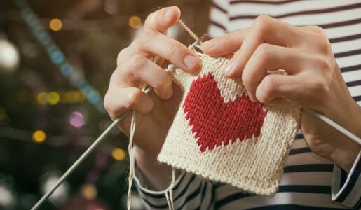 Nahaufnahme von strickenden Händen, die ein rotes Herz auf weißem Strickstück fertigen, vor festlichem Hintergrund. | © svetazi/stock.adobe.com