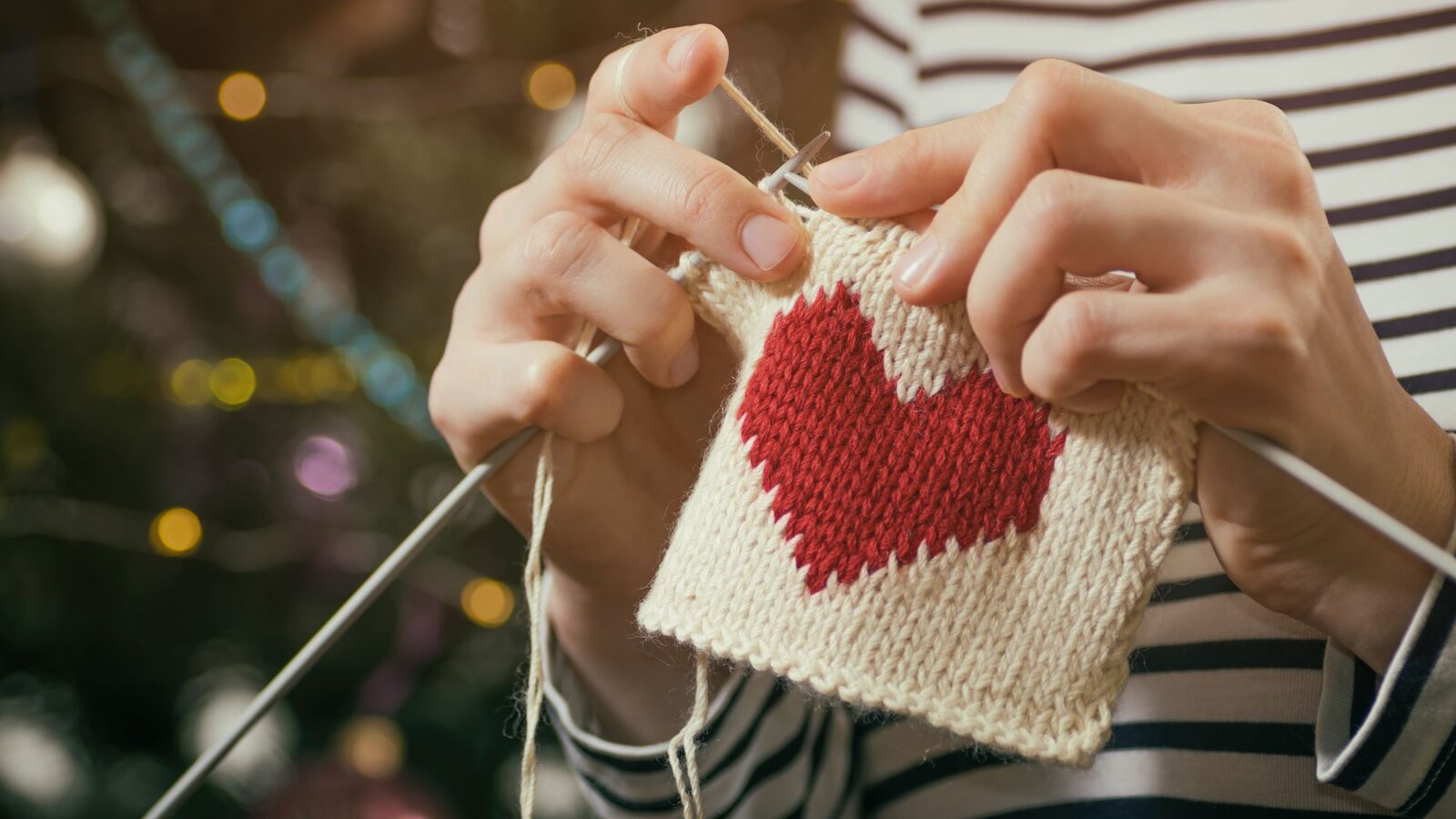 Nahaufnahme von strickenden Händen, die ein rotes Herz auf weißem Strickstück fertigen, vor festlichem Hintergrund. | © svetazi/stock.adobe.com