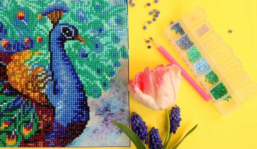 Detailreiches Diamond-Painting eines blauen Pfaus, umgeben von grünen und blauen Strasssteinen, mit Tulpe und Werkzeug auf gelbem Hintergrund. | © Olga/stock.adobe.com