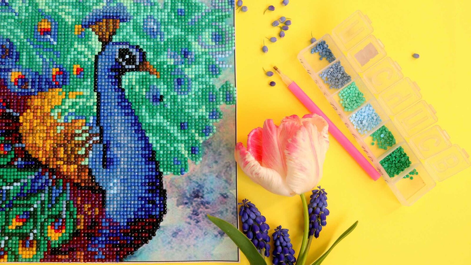 Detailreiches Diamond-Painting eines blauen Pfaus, umgeben von grünen und blauen Strasssteinen, mit Tulpe und Werkzeug auf gelbem Hintergrund. | © Olga/stock.adobe.com