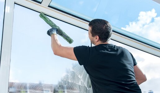 Mann mit schwarzem Shirt steht an großer Fensterscheibe mit Blick in den Garten, er putzt sie mit einem Wasserabzieher, Wetter draußen ist gut und sonnig | © Adobe Stock/pictworks