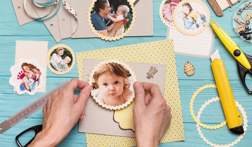 Hände gestalten ein Scrapbook auf blauem Holztisch, dekoriert mit ausgeschnittenen Babyfotos, Bastelpapier, Schere und Cutter. | © Sergii Mostovyi /stock.adobe.com