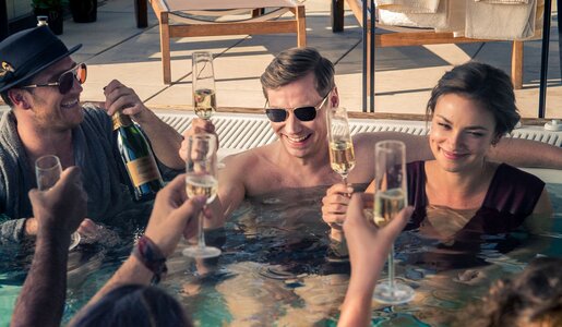 Produktionsbild "Betonrausch": Zwei Männer und eine Frau stoßen im warmen Whirlpool mit Champagner an, umgeben von ausgelassener Atmosphäre und Sonnenlicht. | © © NIK KONIETZNY