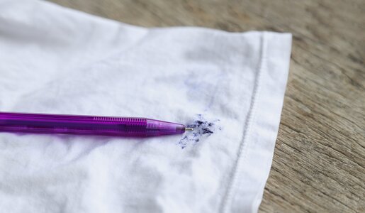 Ein lila Kugelschreiber liegt auf einem weißen Stoff, der mit blauer Tinte befleckt ist, auf einer Holzoberfläche. | © Adobe Stock/ Apiwat