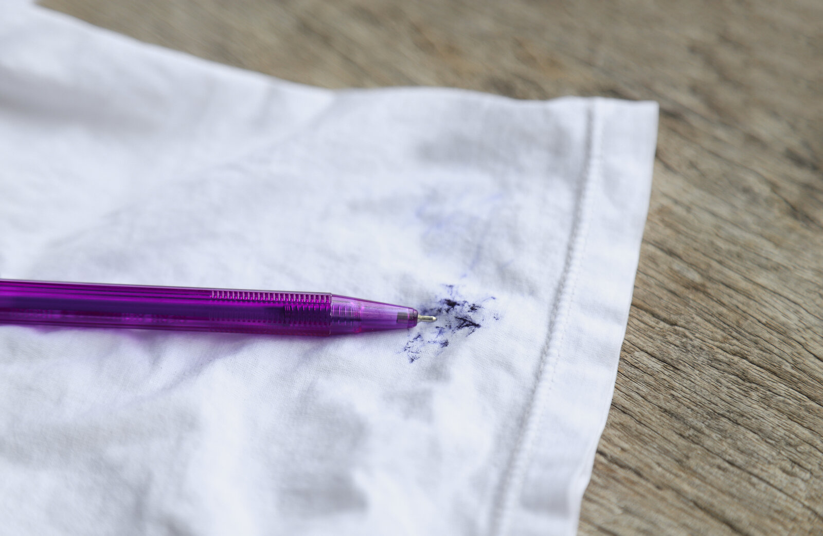 Ein lila Kugelschreiber liegt auf einem weißen Stoff, der mit blauer Tinte befleckt ist, auf einer Holzoberfläche. | © Adobe Stock/ Apiwat