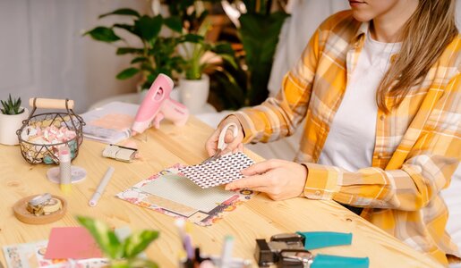 Eine Frau mit blondbraunen Haaren und kariertem Hemd bastelt an einem Holztisch mit Schere, Papier und Scrapbooking-Material. | © Anna/stock.adobe.com