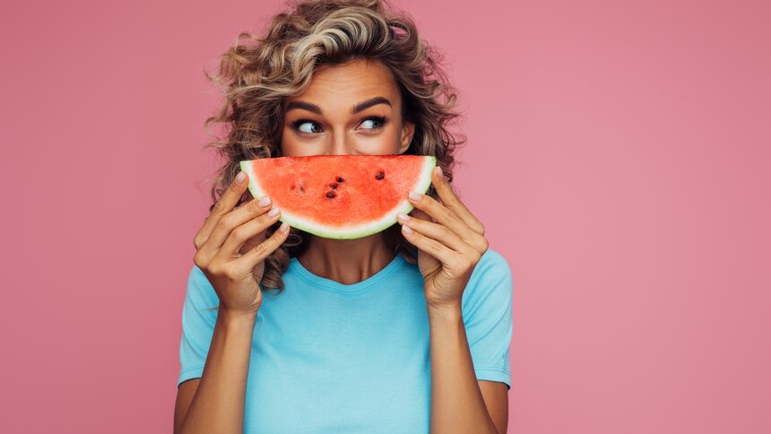Eine junge Frau mit blonden Locken hält sich ein Stück Wassermelone vor das Gesicht. Der Hintergrund ist komplett pink eingefärbt und die Frau trägt ein hellblaues T-Shirt. | © Getty Images / CoffeeAndMilk