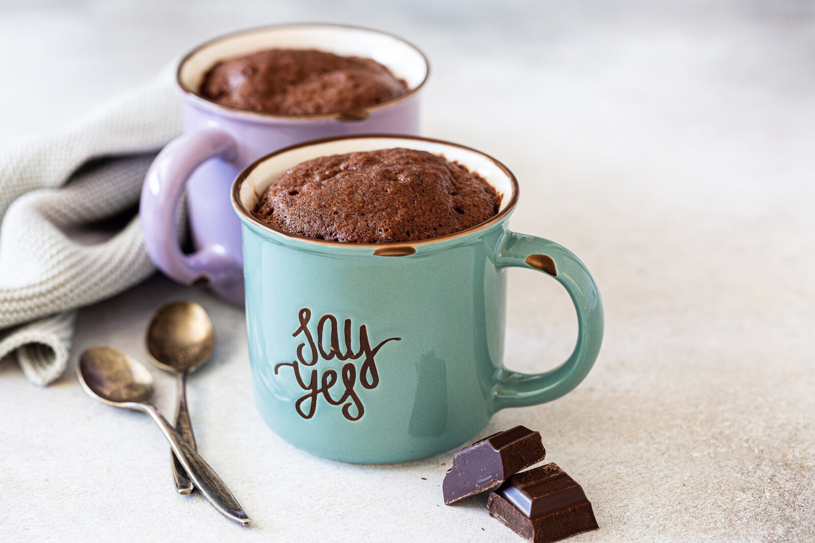 Zwei Tassen mit fertigem Schoko-Küchlein. Eine Tasse ist blau, die andere lila. Auf den Tassen steht "say yes" und neben den Tasser liegen zwei Teelöffel, zwei Stücke Schokolade und ein Geschirrtuch. | © Getty Images / Iryna Melnyk