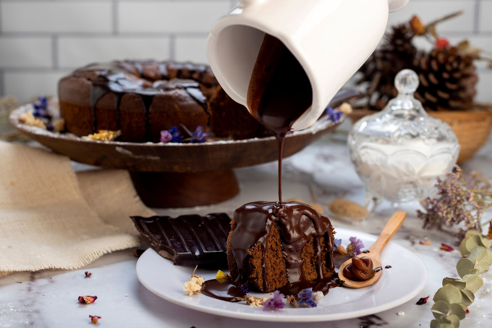 Schokokuchen auf einer Kuchenplatte, im Vordergrund ein Stück Schokokuchen, der von einer Hand mit flüssiger Schokolade überzogen wird.  | © Getty Images / Wirestock