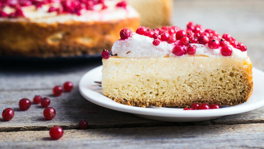Ein Stück Johannisbeer-Torte liegt auf einem weißen Teller. Im Hintergrund und etwas unscharf sieht man die ganze Torte. | © Getty Images / ollinka
