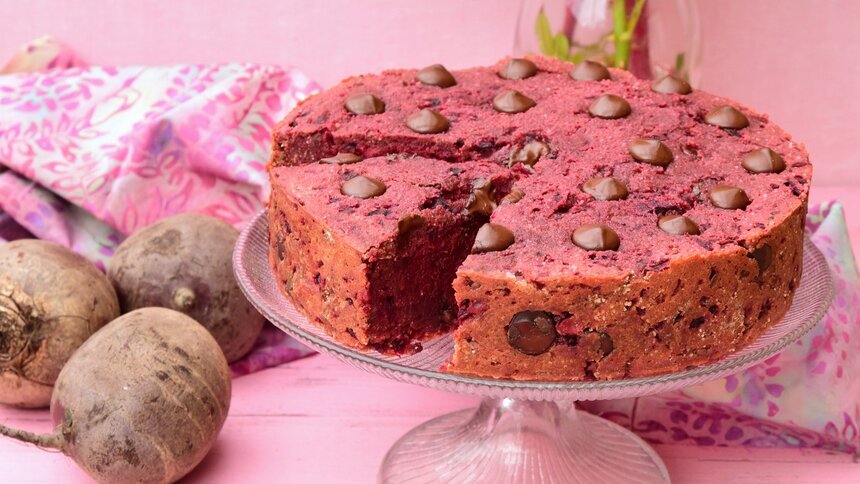 Rote-Bete-Kuchen steht auf einem Kuchenteller. Der Hinterggrund ist rosa und neben dem Kuchen liegt frische Rote-Bete. | © Getty Images / AmalliaEka