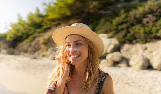 Blonde und junge Frau trägt einen Hut und ist am Strand. Sie trägt sich Streifen von Sonnencreme auf die Wangen und lächelt dabei. | © Getty Images / AleksandarNakic
