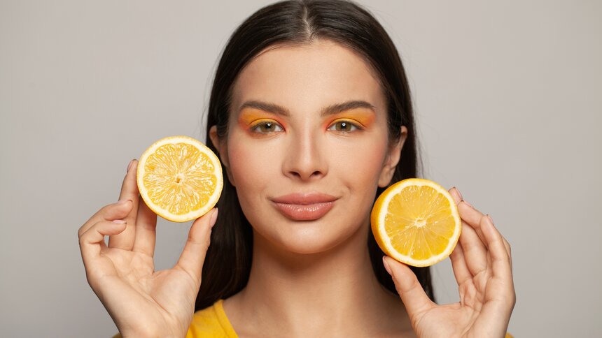 Eine junge Frau mit dunklen Haaren und orangem Makeup hält zwei Orangenschalen in den Händen. | © Getty Images / JNemchinova