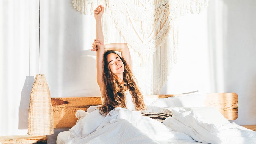 Junge Frau mit dunklen langen Haaren reckt und streckt sich im Bett. | © Getty Images / Maria Korneeva