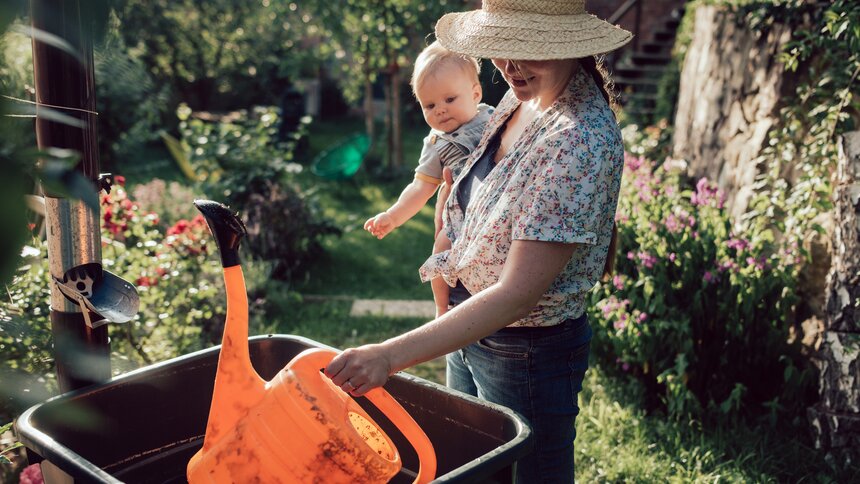 Eine junge Frau hält ein Baby auf dem Arm und tunkt eine Gießkanne in einen vollen Regenwassertank im Garten. | © Getty Images / Guido Mieth