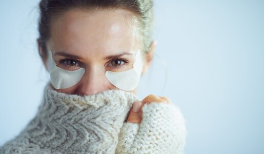 Eine Frau mittleren Alters trägt einen dicken Wollpullover und trägt zusätzlich unter den Augen feuchtigkeitsspendende Augen-Pads. | © Getty Images / CentralITAlliance