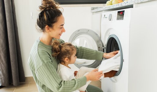 Frau mit Baby auf dem Schoss befüllt Waschmaschine mit Wäsche. | © Getty Images / Anastasiia Krivenok; 