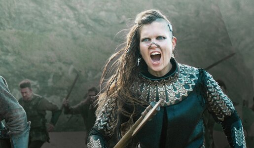 Junge Frau mit wehendem langen Haar in altertümlicher Rüstung mit gezogenem Schwert stürmt in einer Szene der Serie "Cursed" zusammen mit Männern. im Hintergrund ein Berg. | © Netflix