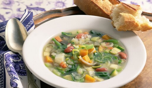 Gemüsesuppe angerichtet in weißem Suppenteller, daneben Baguette-Scheiben und ein Löffel. | © Getty Images / 	Brian Hagiwara