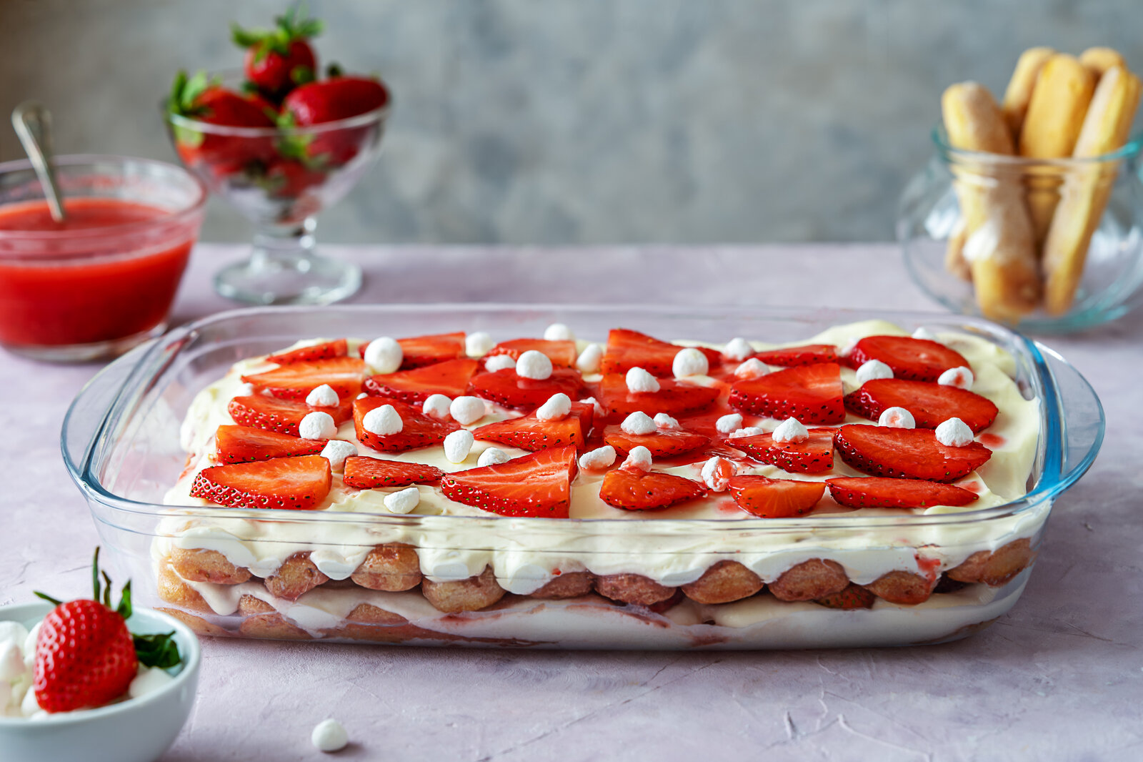 Erdbeer-Tiramisu, hergestellt im Thermomix in einer Glasform auf einem Tisch stehend. Zu sehen sind außerdem die enthaltenen Löffelbiskuits, frische Erdbeeren und Erdbeersoße. | © Getty Images / Tetiana Zavgorodnia
