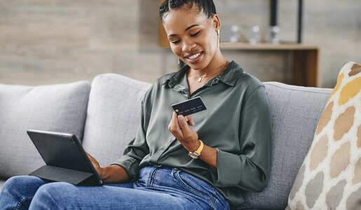 Frau in grüner Bluse und blauer Jeans sitzt lächelnd auf einem Sofa und hält eine Kreditkarte in der Hand, während sie ein Tablet benutzt. | © Getty Images/Charday Penn