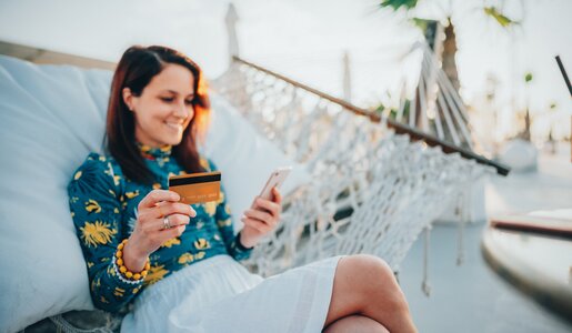 Eine junge Frau mit braunen Haaren sitzt lächelnd in einer Hängematte, hält eine Kreditkarte und ein Smartphone. | © Getty Images/martin-dm