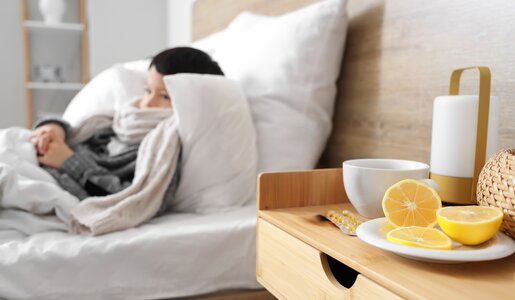 Nahaufnahme eines Nachttischs mit Tee und Zitronenscheiben, im Hintergrund ein Kind mit Schal im Bett. | © Adobe Stock/ Pixel-Shot
