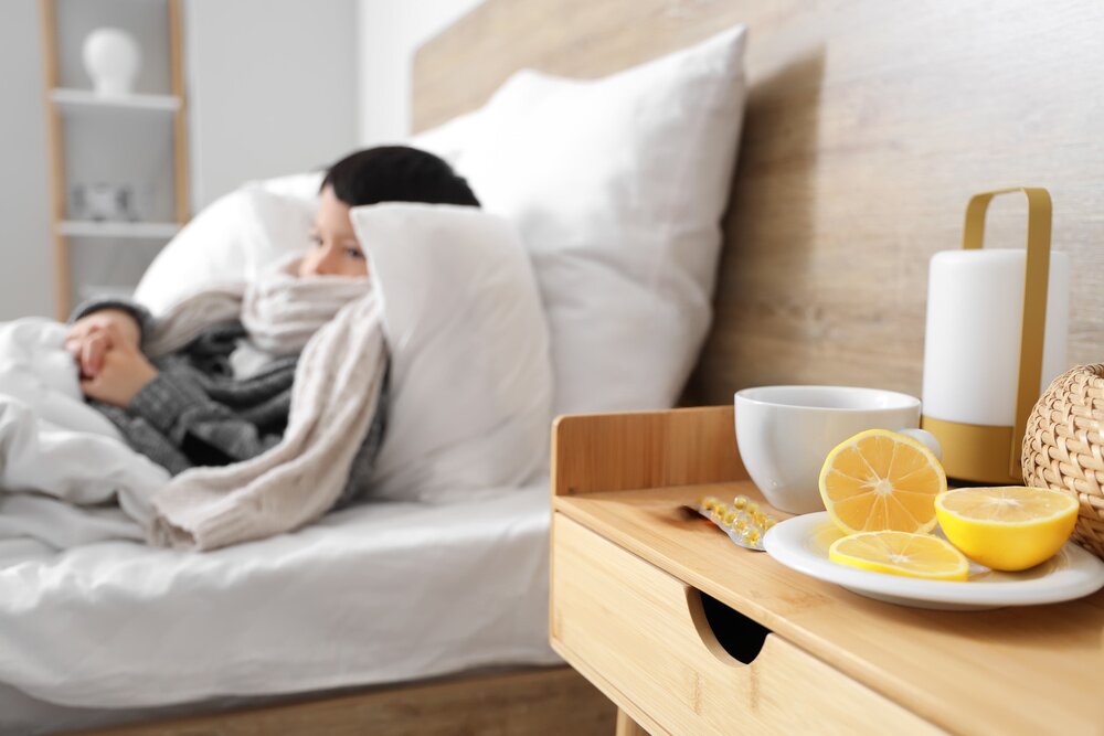 Nahaufnahme eines Nachttischs mit Tee und Zitronenscheiben, im Hintergrund ein Kind mit Schal im Bett. | © Adobe Stock/ Pixel-Shot