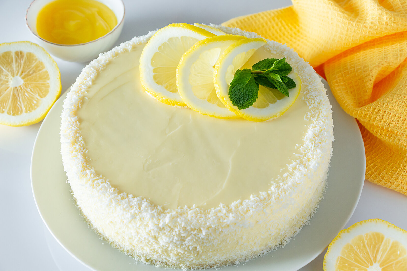 Angeschnittener Kokos-Käsekuchen auf einem weißen Teler, daneben und obendrauf frische Zitronenscheiben und ein Minzblatt. | © Getty Images / Galina Atroshchenko
