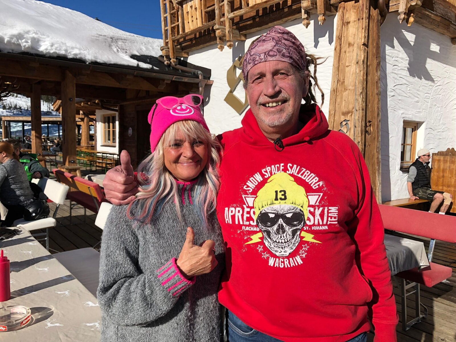 Ein Mann in rotem Hoodie und eine Frau mit rosa Mütze stehen bei Sonnenschein vor einer rustikalen Skihütte. | © RTL/99pro media