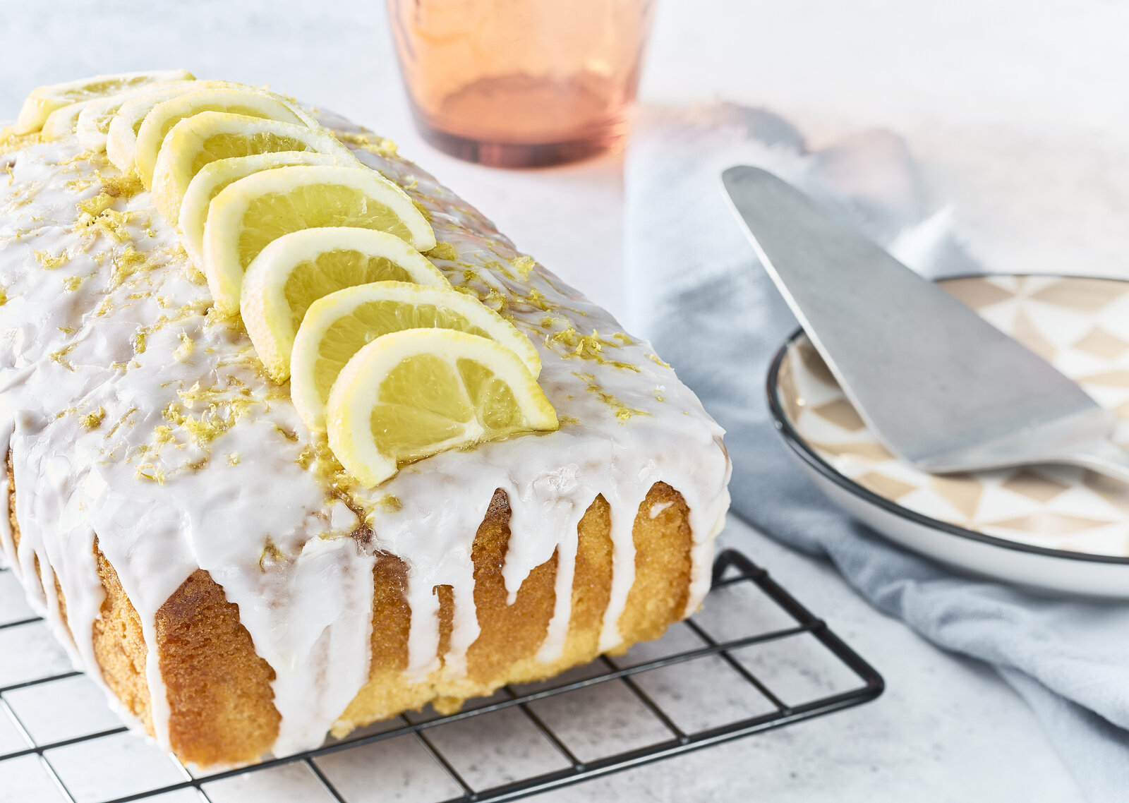 Angeschnittener Zitronenkuchen mit Joghurtguss auf einem Abkühlgitter, daneben Tortenheber auf einem Teller. | © Getty Images / Tim Young Photography