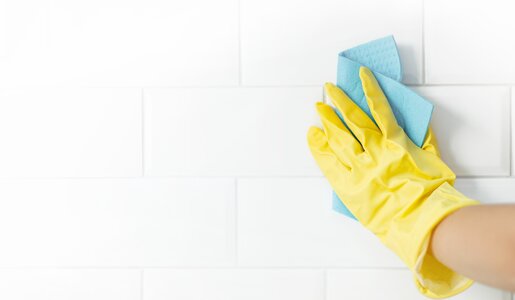 Eine Hand in gelbem Gummihandschuh wischt mit einem blauen Tuch eine weiße geflieste Wand. | © Adobe Stock/ Алина Битта