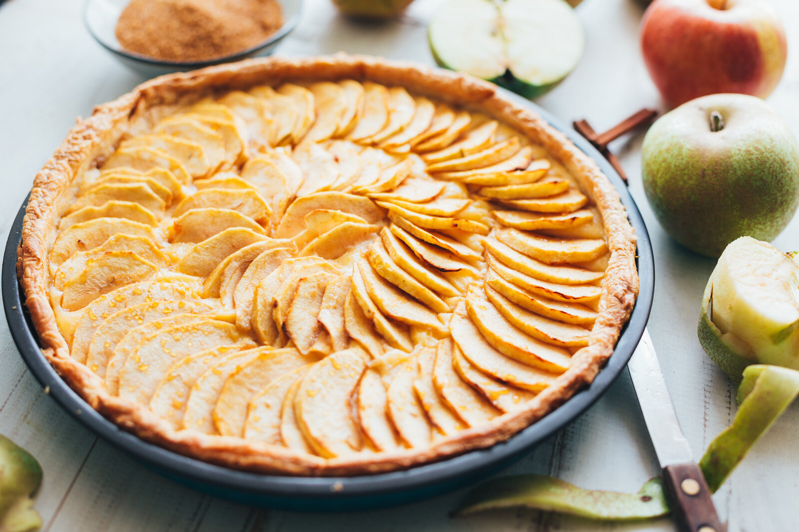 Apfelkuchen in Backform auf dem Tischstehend, Apfelstücke auf Oberfläche drapiert und neben dem Kuchen liegen weitere frische Äpfel. | © Adobe Stock / asife