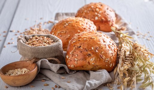 drei Haferbrötchen auf einer Holzunterlage daneben Weizenzweige und Körner. | © Adobe Stock / shaiith