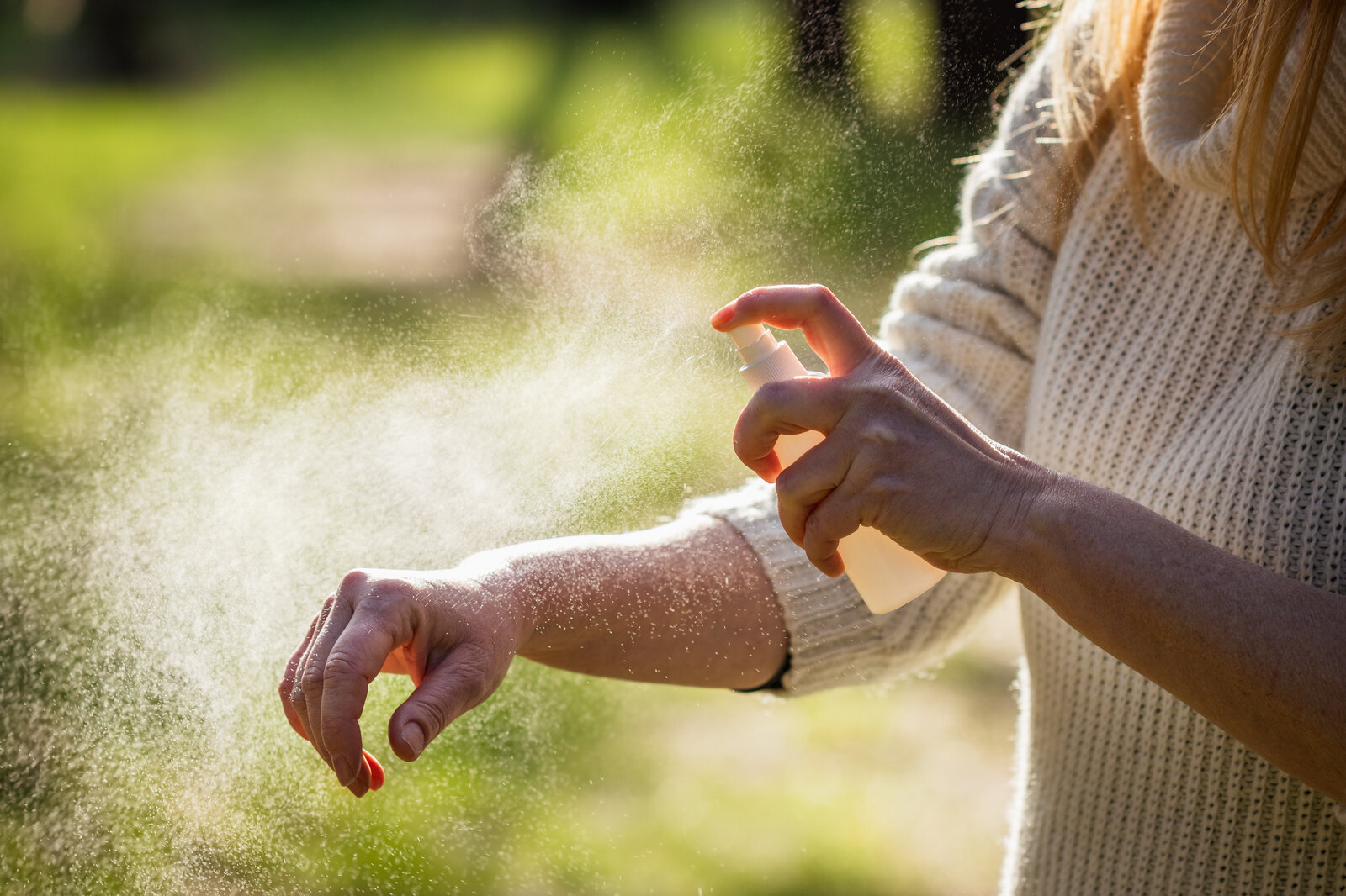 Eine Frau in weißem Strickpullover sprüht sich im Sonnenschein Insektenspray auf den Unterarm. | © Adobe Stock/ encierro