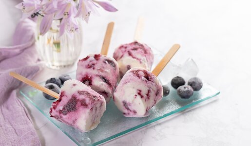 Vier kleine Joghurteis am Stiel mit Blaubeeren, daneben frische Blaubeeren in fliederfarbener Umgebung. | © Getty Images / yumehana