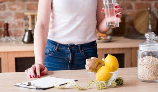 Eine Frau in weißem Top und Jeans hält ein Glas Wasser in der Küche, neben ihr liegen Zitronen, Ingwer und ein Maßband auf dem Tisch. | © Adobe Stock/ golubovy