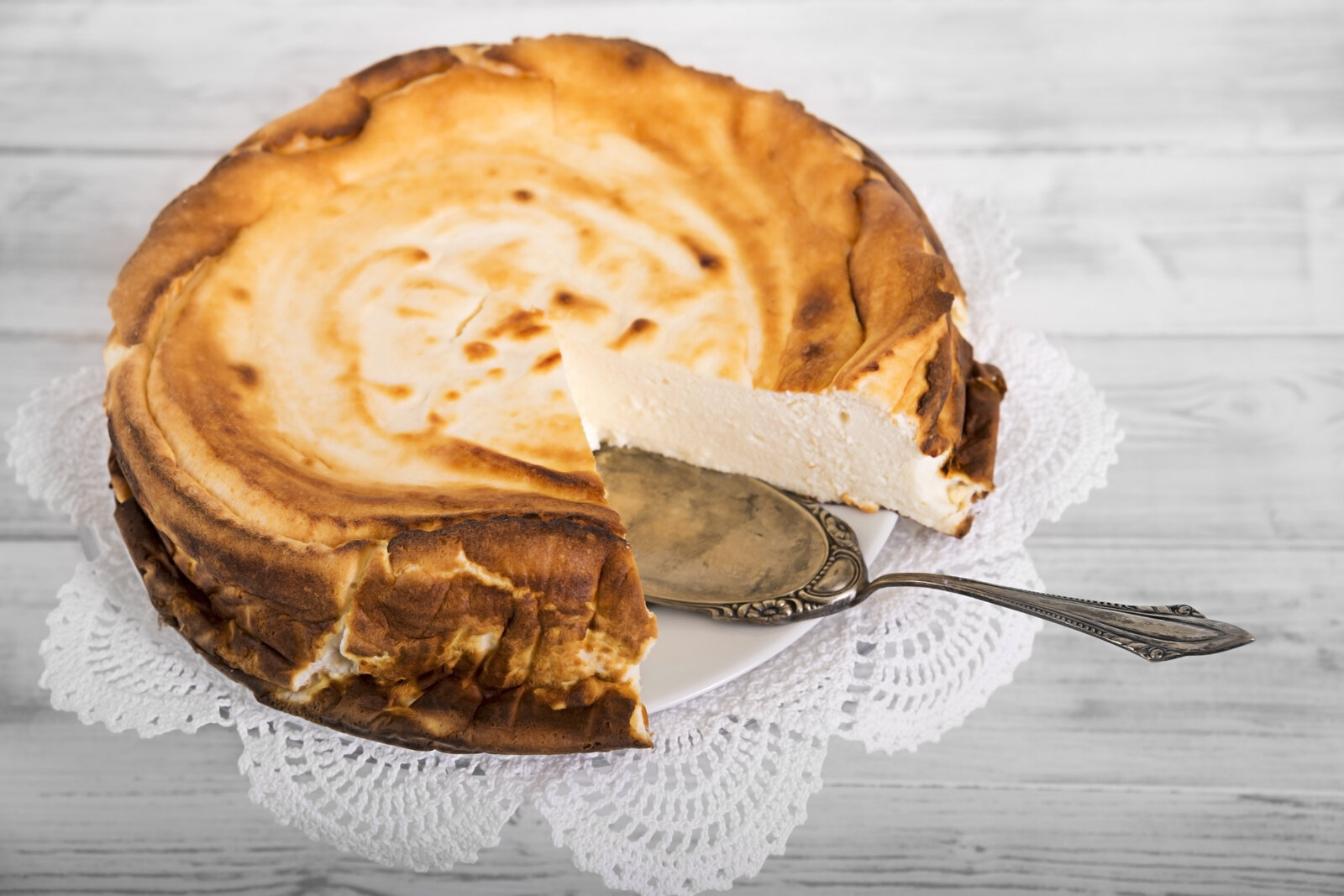 Angeschnittener Käsekuchen auf einem weißen Tortenboden, daneben ein silberner Tortenheber. | © Getty Images / Westend61