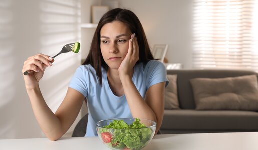 Junge Frau in hellblauem T-Shirt betrachtet gelangweilt eine Gurkenscheibe auf einer Gabel vor einer Schüssel Salat im hellen Wohnzimmer. | © Adobe Stock/ New Africa