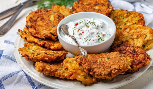 Nebeneinander angerichtete Zucchini-Karotten-Puffer auf einem Teller, in der Mitte eine kleine Schale mit weißem Dip. | © Getty Images / 	SMarina
