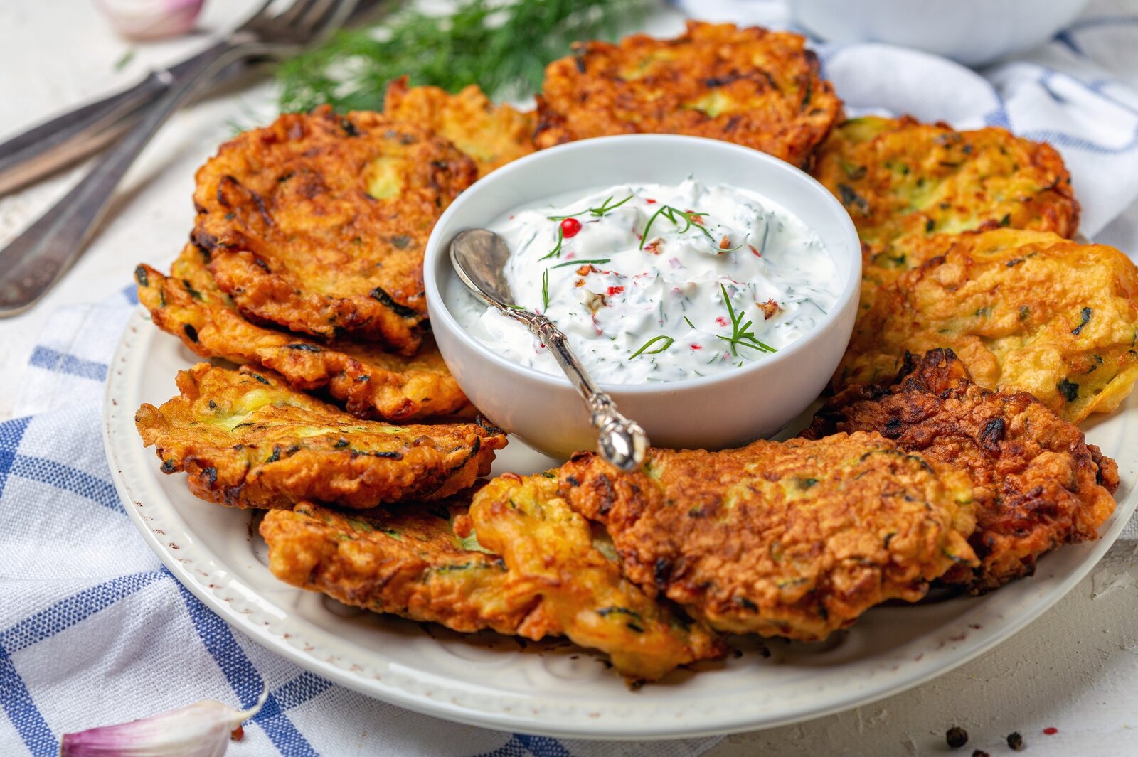 Nebeneinander angerichtete Zucchini-Karotten-Puffer auf einem Teller, in der Mitte eine kleine Schale mit weißem Dip. | © Getty Images / 	SMarina