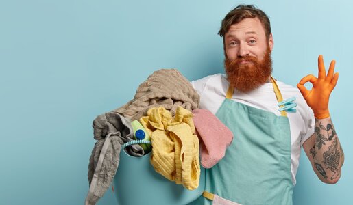 Ein bärtiger, tätowierter Mann in mintgrüner Schürze zeigt lächelnd das "OK"-Zeichen und hält einen Eimer voller bunter Wäsche. | © Adobe Stock/ wayhome.studio