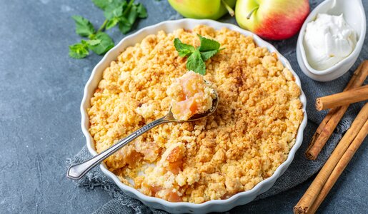 Apple Crumble in Auflaufform, mit einem Löffel drinnen stecken, daneben frische Äpfel, Eis und frische Minze. | © Adobe Stock / sriba3