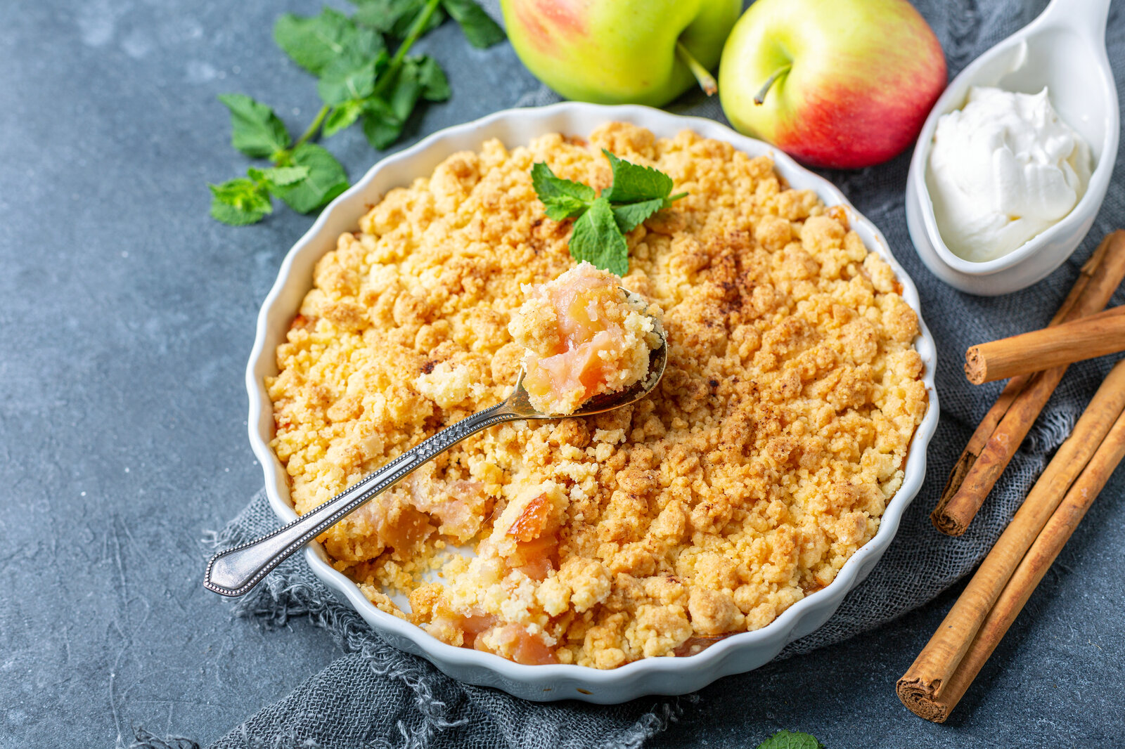 Apple Crumble in Auflaufform, mit einem Löffel drinnen stecken, daneben frische Äpfel, Eis und frische Minze. | © Adobe Stock / sriba3