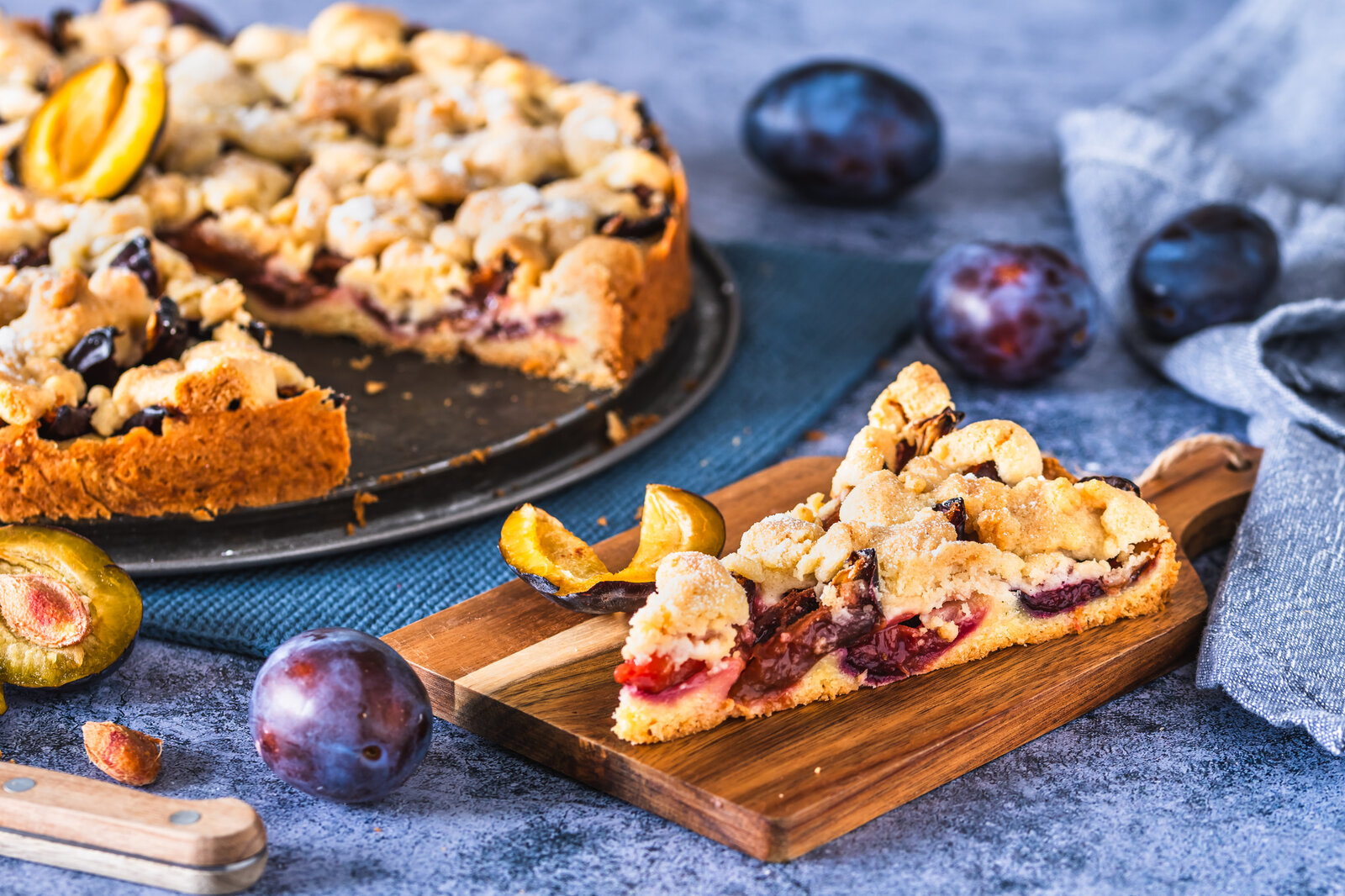 Angeschnittener Pflaumenkuchen auf einem Kuchenteller, im Vordergrund ein Stück des Kuchens auf einem weiteren Teller. | © Getty Images / Daniela Baumann